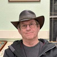 Man wearing a brown hat and glasses, seated indoors against a light green wall.