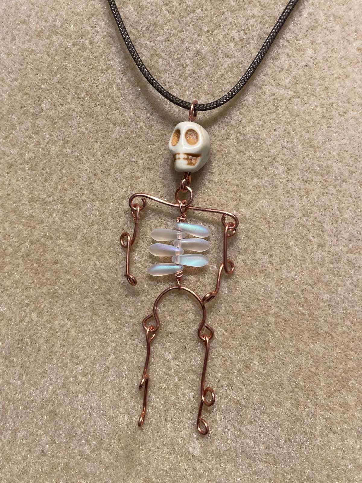 A handmade wire-wrapped skeleton pendant on a dark beaded chain, featuring a white skull bead and iridescent ribs.