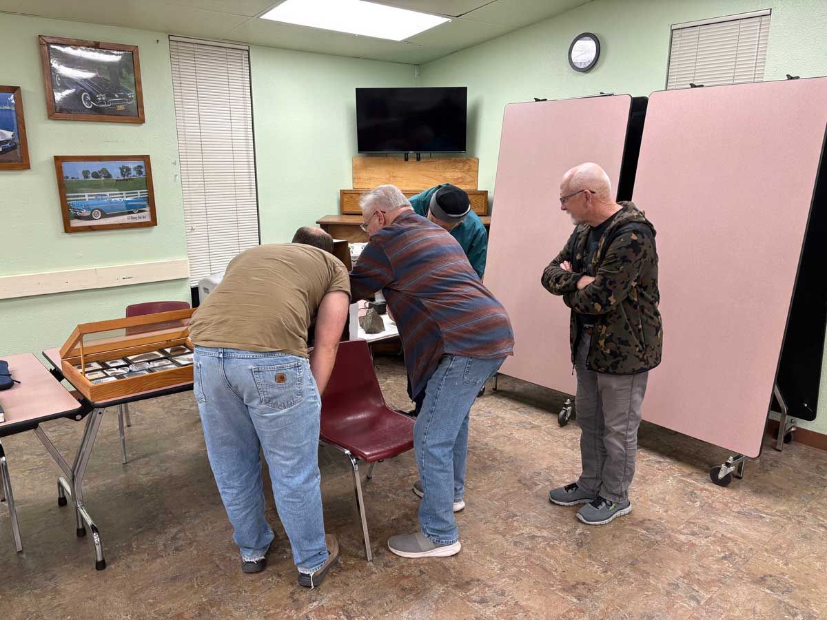 Four individuals gather in a community room, examining items on a table and chair near a large display board.