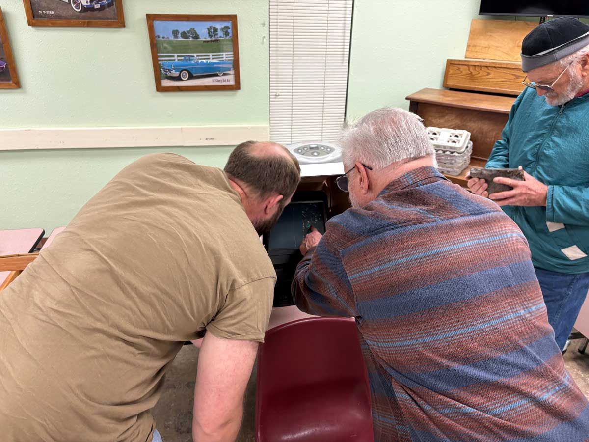 Three people examine an object on a low table in a room decorated with framed car photos.