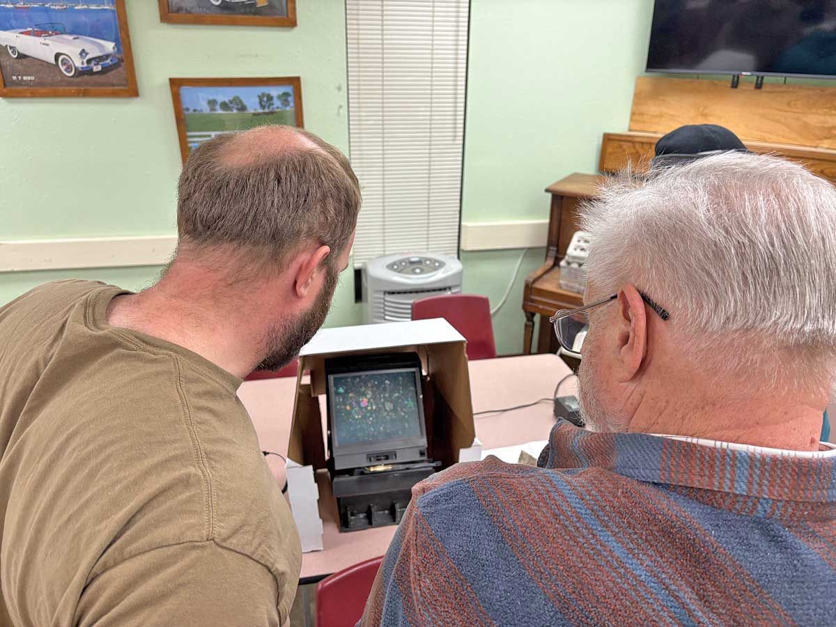 Two people look into a tabletop light box displaying a slide in a room decorated with framed car art.