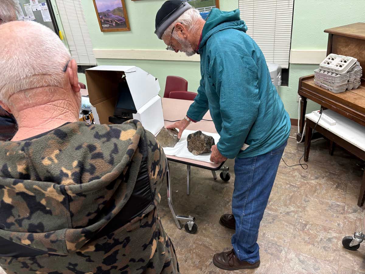 Two people examine a dark, porous rock placed on a white paper-covered table in an indoor room.