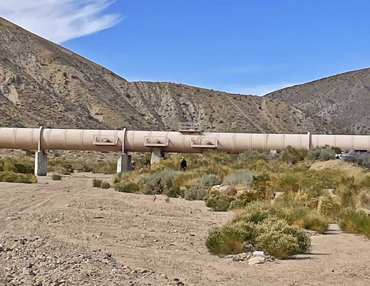 Rusty pipeline crossing a dry desert landscape with scrub and rocky hills under a blue sky