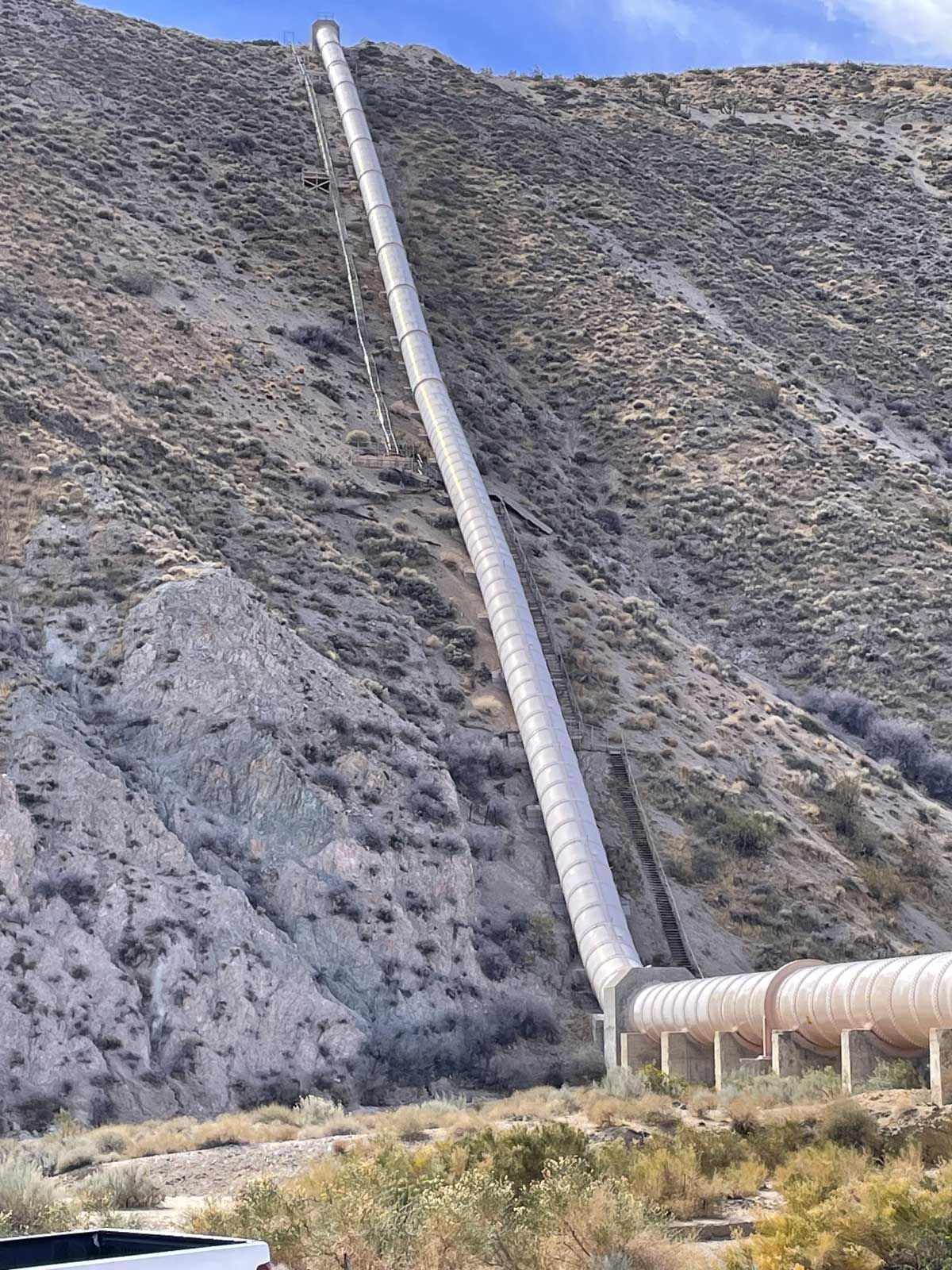 Long white pipeline climbs a rocky hillside beside a smaller pipeline at the base.