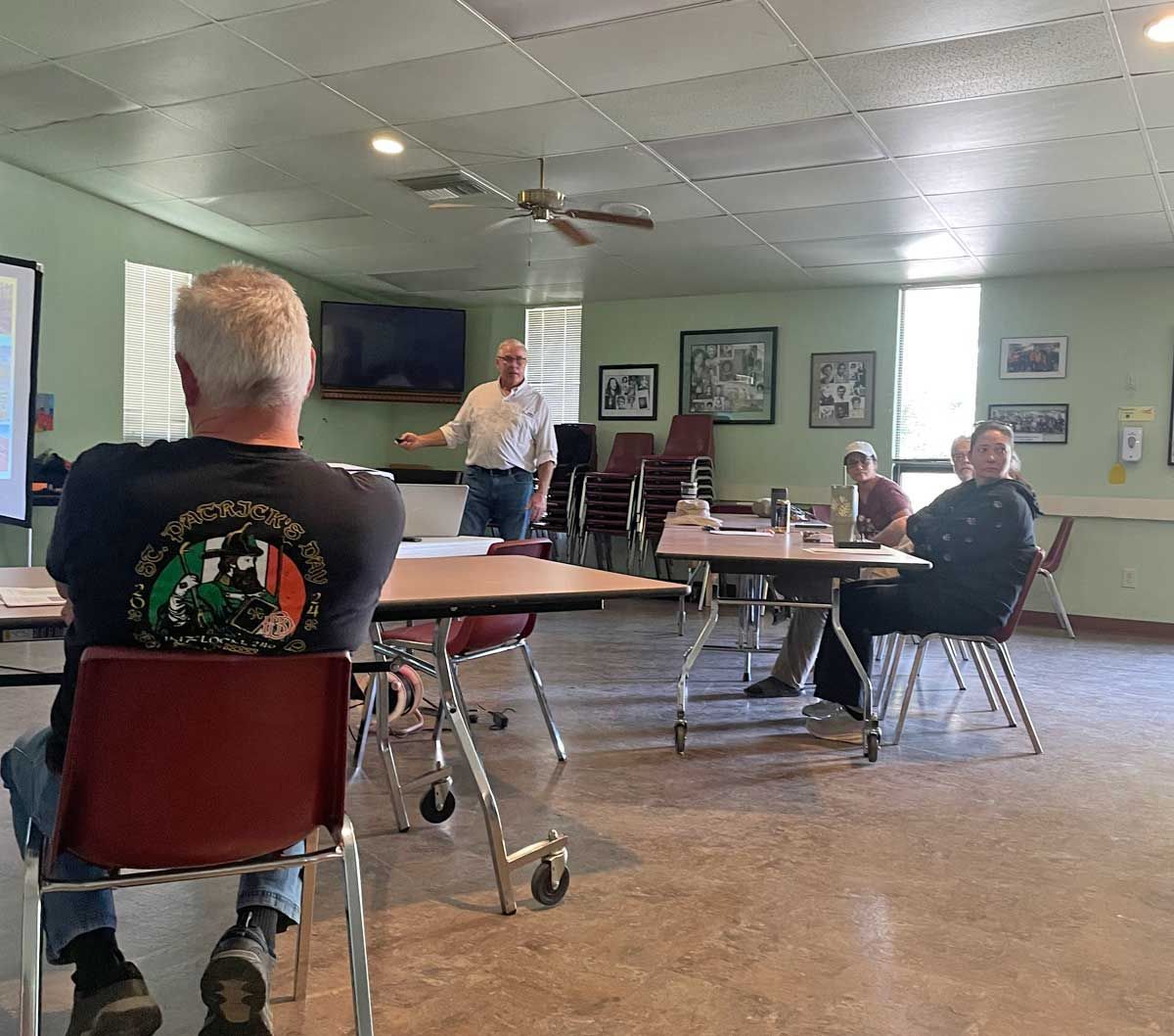 People seated in a community hall while a man speaks near the front wall-mounted TV.
