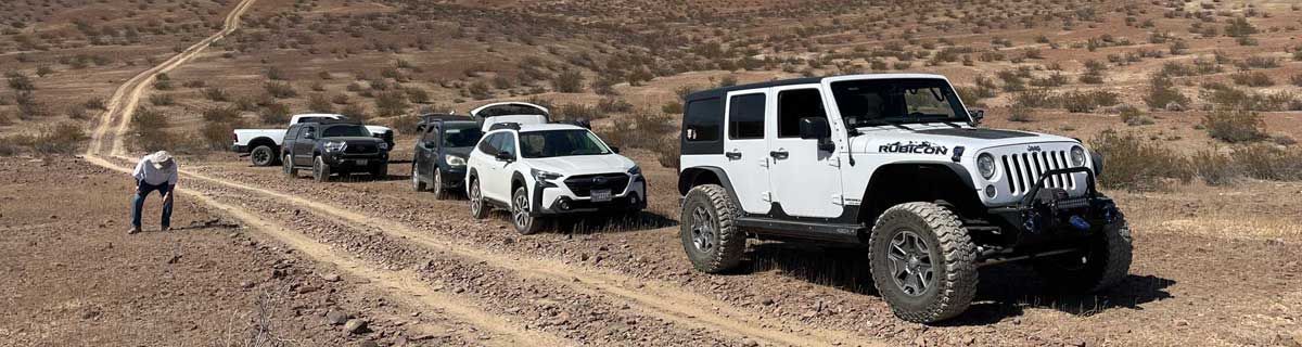 Three off-road jeeps on a dirt trail in a rocky desert, with a person walking nearby.
