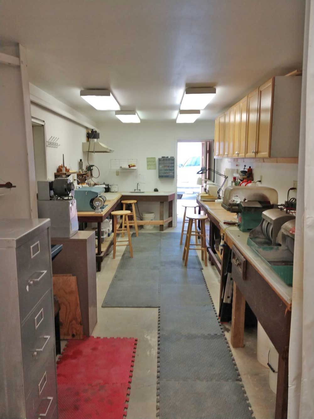 Narrow workshop kitchen with stainless counters, wooden cabinets, stools, and fluorescent lights.