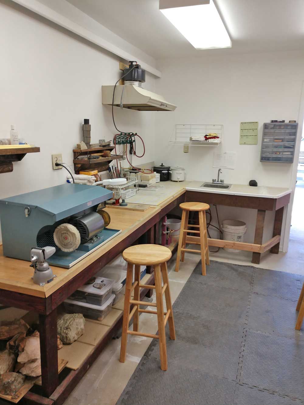 Workshop interior with workbenches, stools, tools, and overhead lights on a tiled floor.