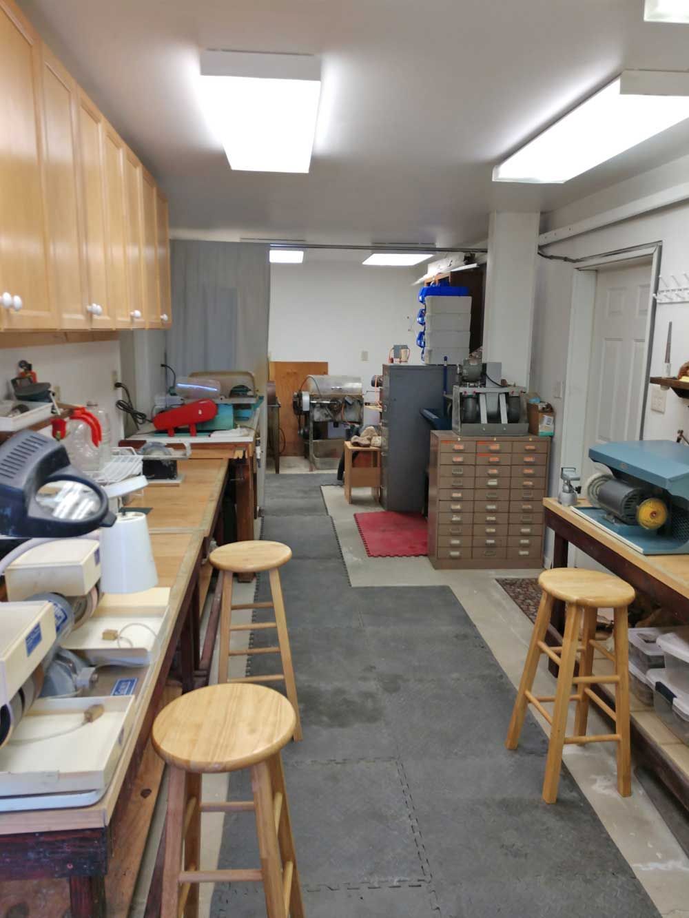 Workshop with workbenches, stools, cabinets, and tools along a narrow aisle under bright fluorescent lights.
