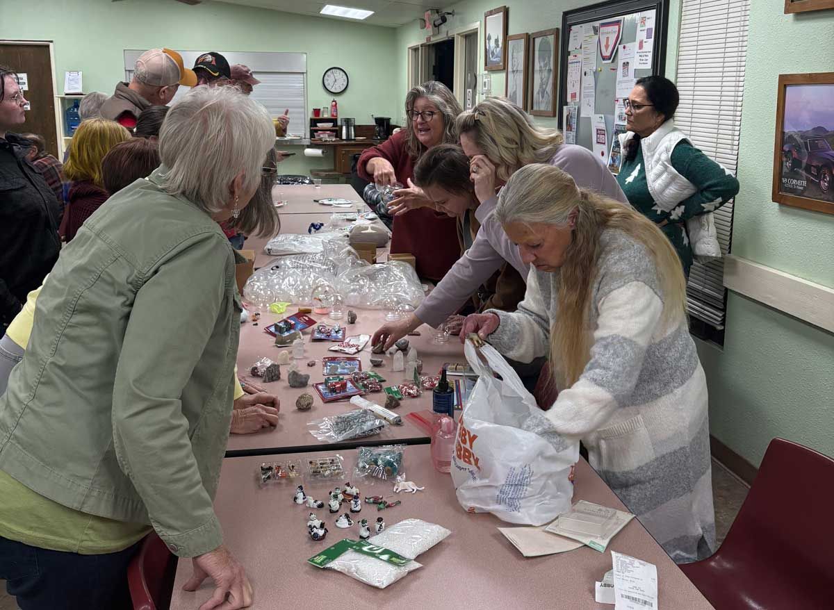People gather around a long table to sort small craft items, plastic bags, and supplies in a community room.