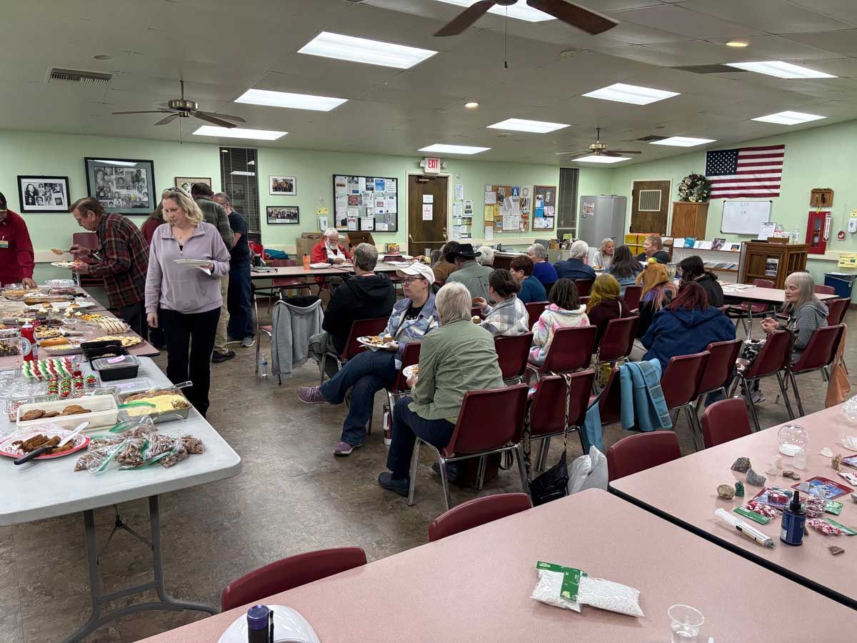 People gather in a community hall with tables filled with food, social interaction, and an American flag on the wall.