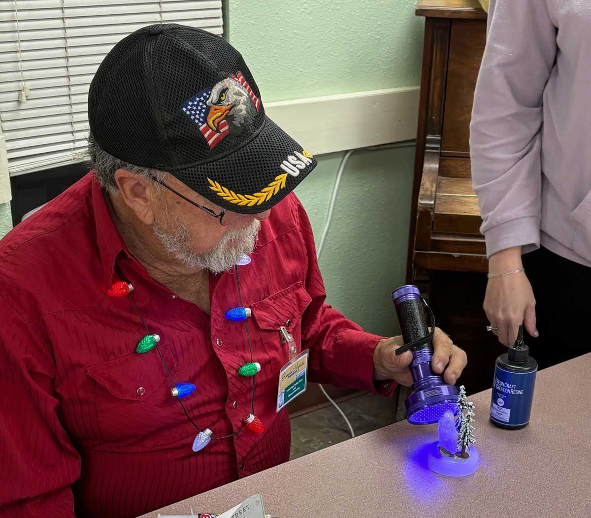A person wearing a red shirt with festive lights uses a purple UV flashlight on a small object on a table.