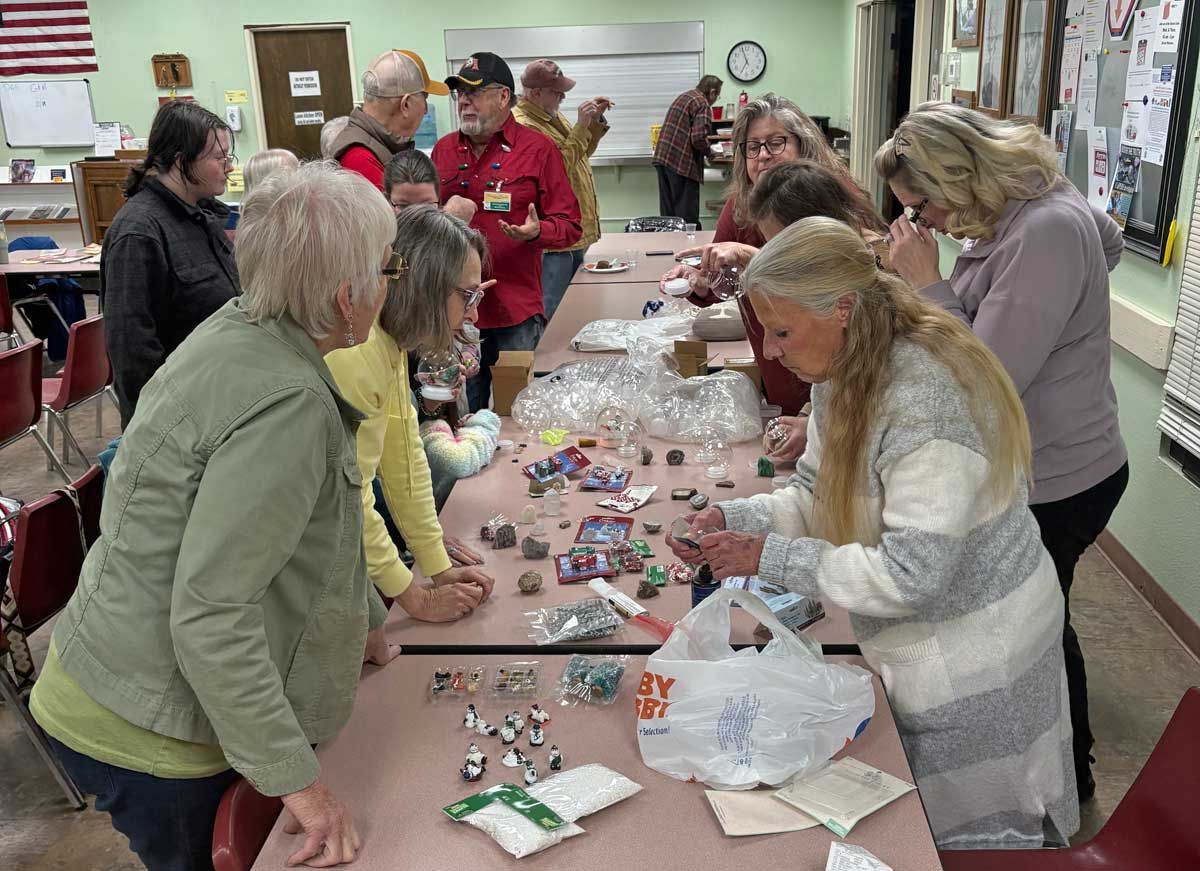 A group of people standing around a table in a community center, engaged in organizing and sorting small craft supplies.