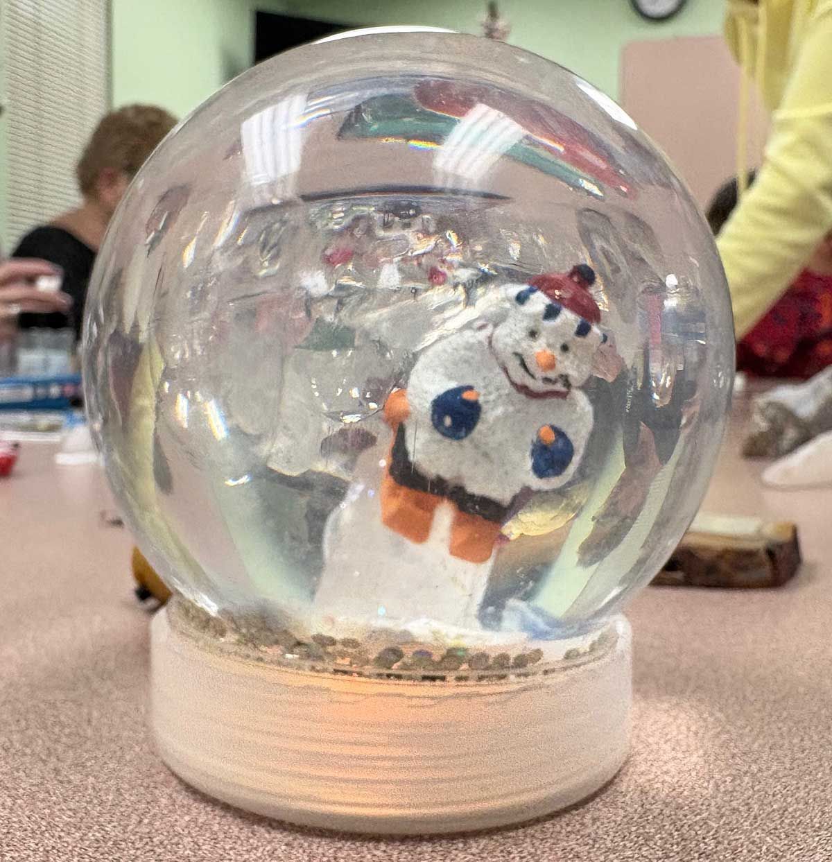 A snowman figure with a red hat and orange mittens inside a glass snow globe on a table.