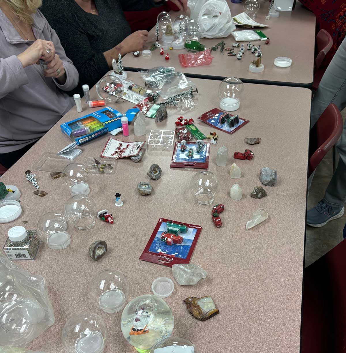 People assemble snow globes on a table scattered with craft supplies, miniature figurines, and various project materials.