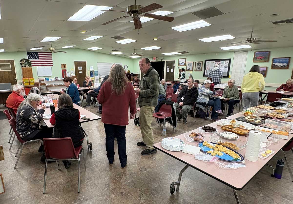 People gather in a community hall for a meal, with tables filled with food and attendees interacting throughout the room.