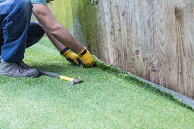Person installing green turf along a wooden fence with a utility knife and gloves