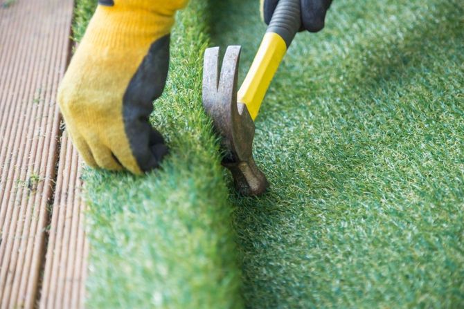 Hands using pliers to pull up artificial grass beside a wooden deck.