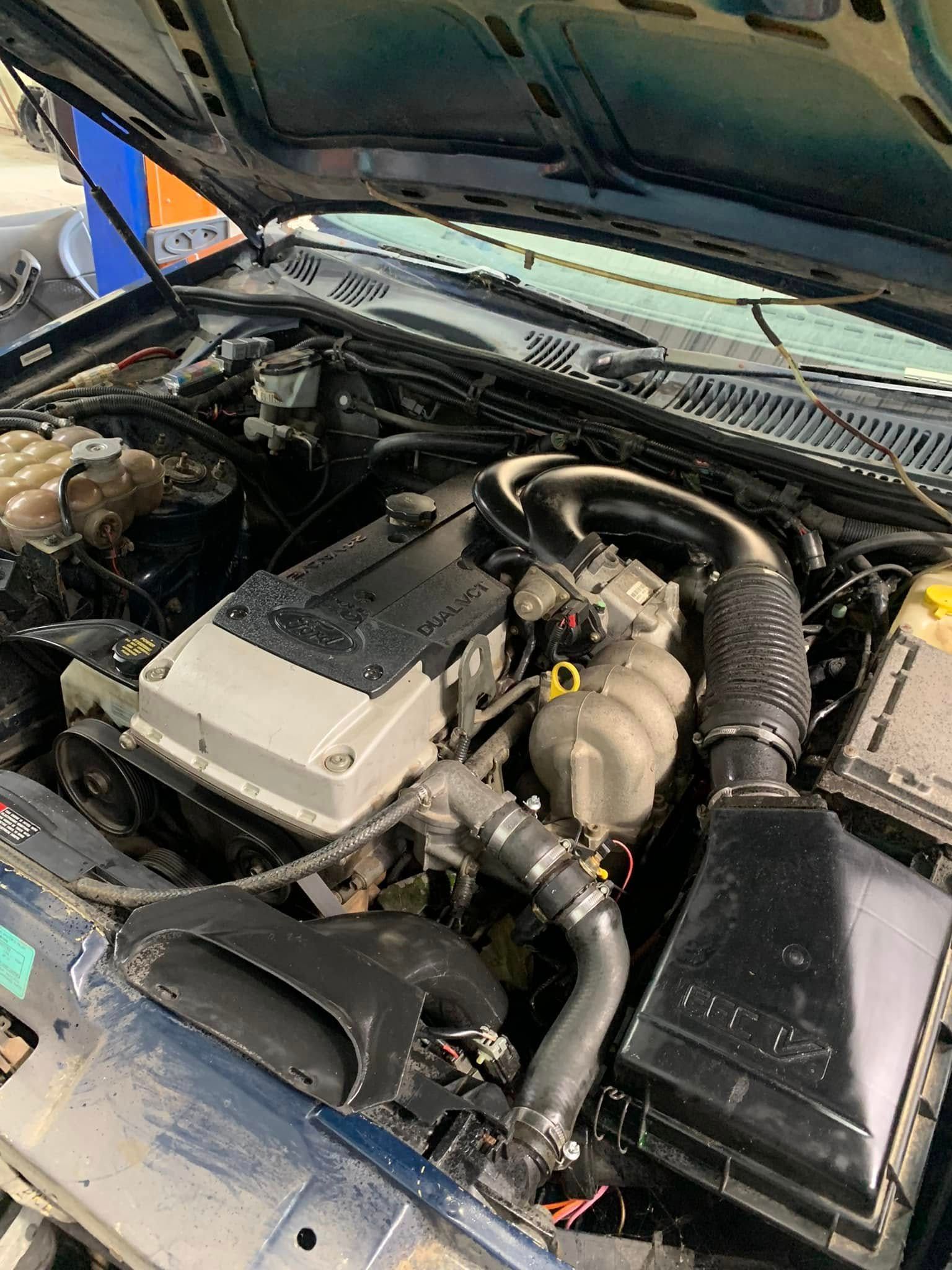 Car engine bay with hood open, showing hoses, air box, and battery components.