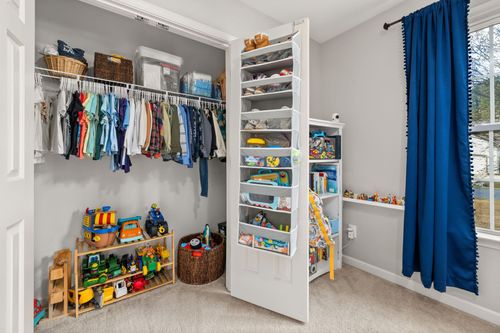 Bright children's closet with hanging clothes, shelves, toys, and a blue curtain by a window.