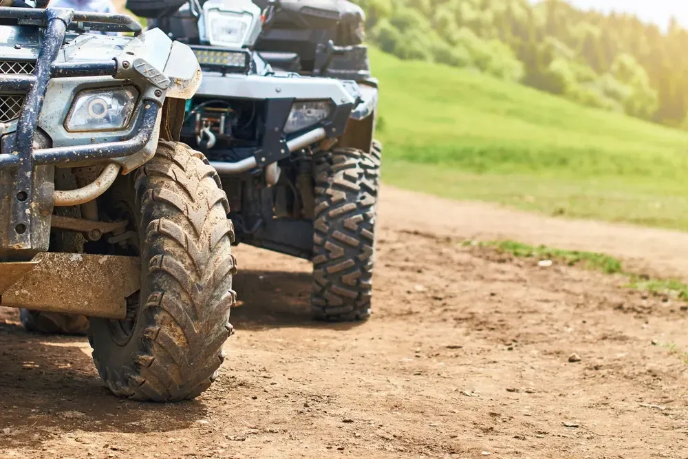 Two all-terrain vehicles (ATVs) parked on a dirt path, tires covered in mud, with a grassy hill in the background. — Jack Too You Mobile Servicing and Repairs In Yandina, QLD