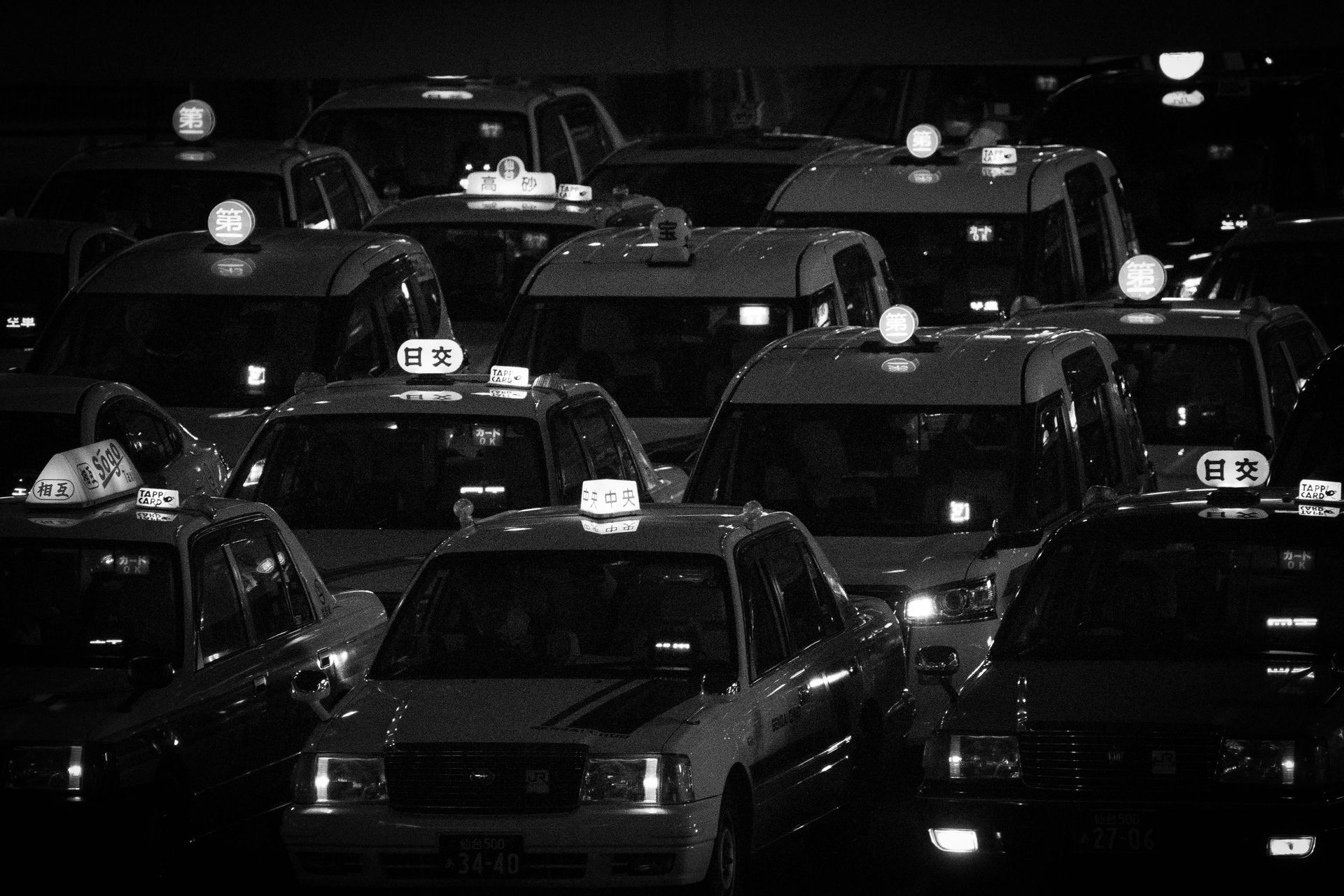 Black and white photo of many Japanese taxis lined up at night, illuminated by headlights and roof lights.