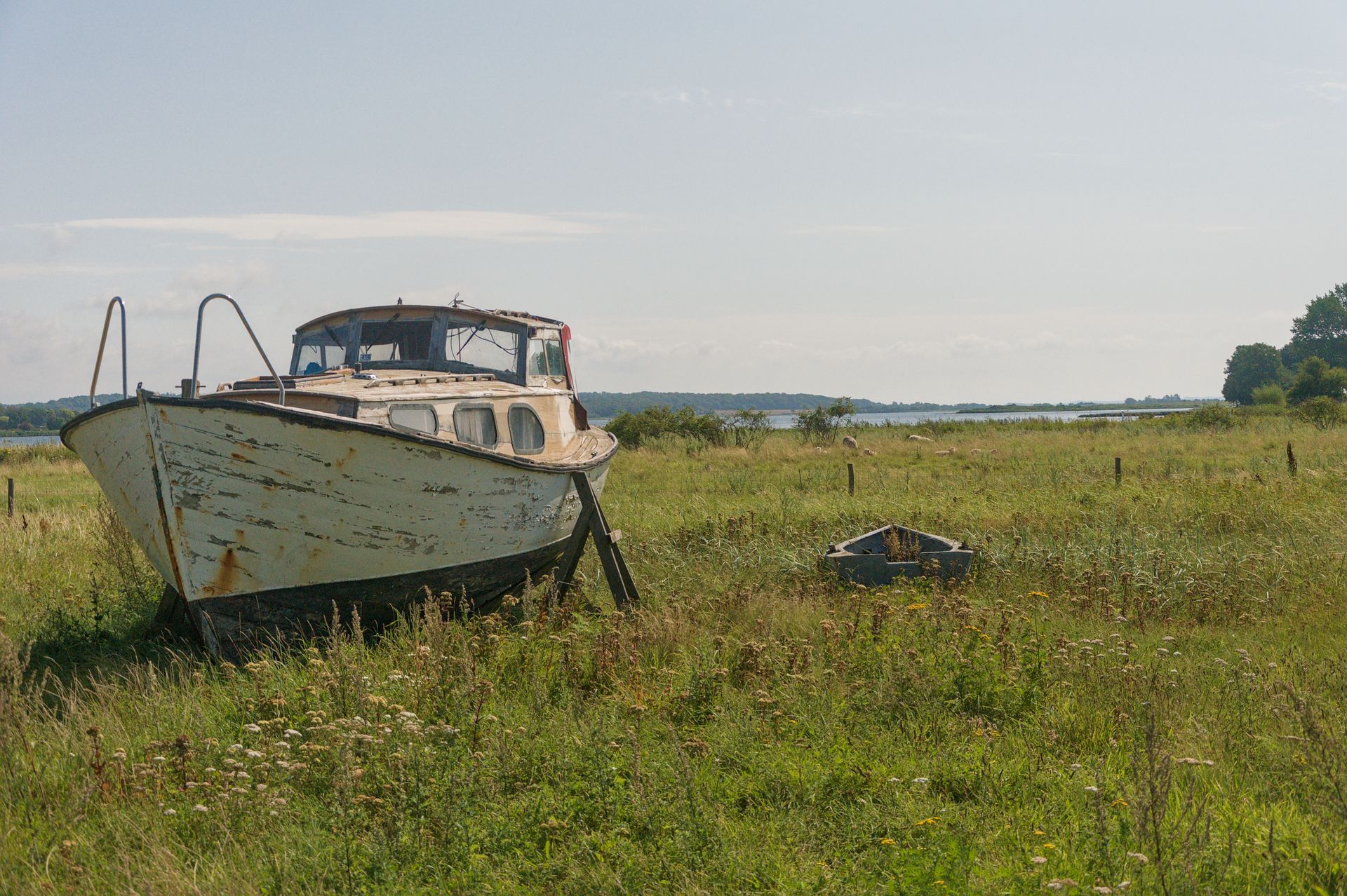Een oude witte boot, gestrand in een grasveld onder een heldere hemel, met in de verte de horizon en bomen.