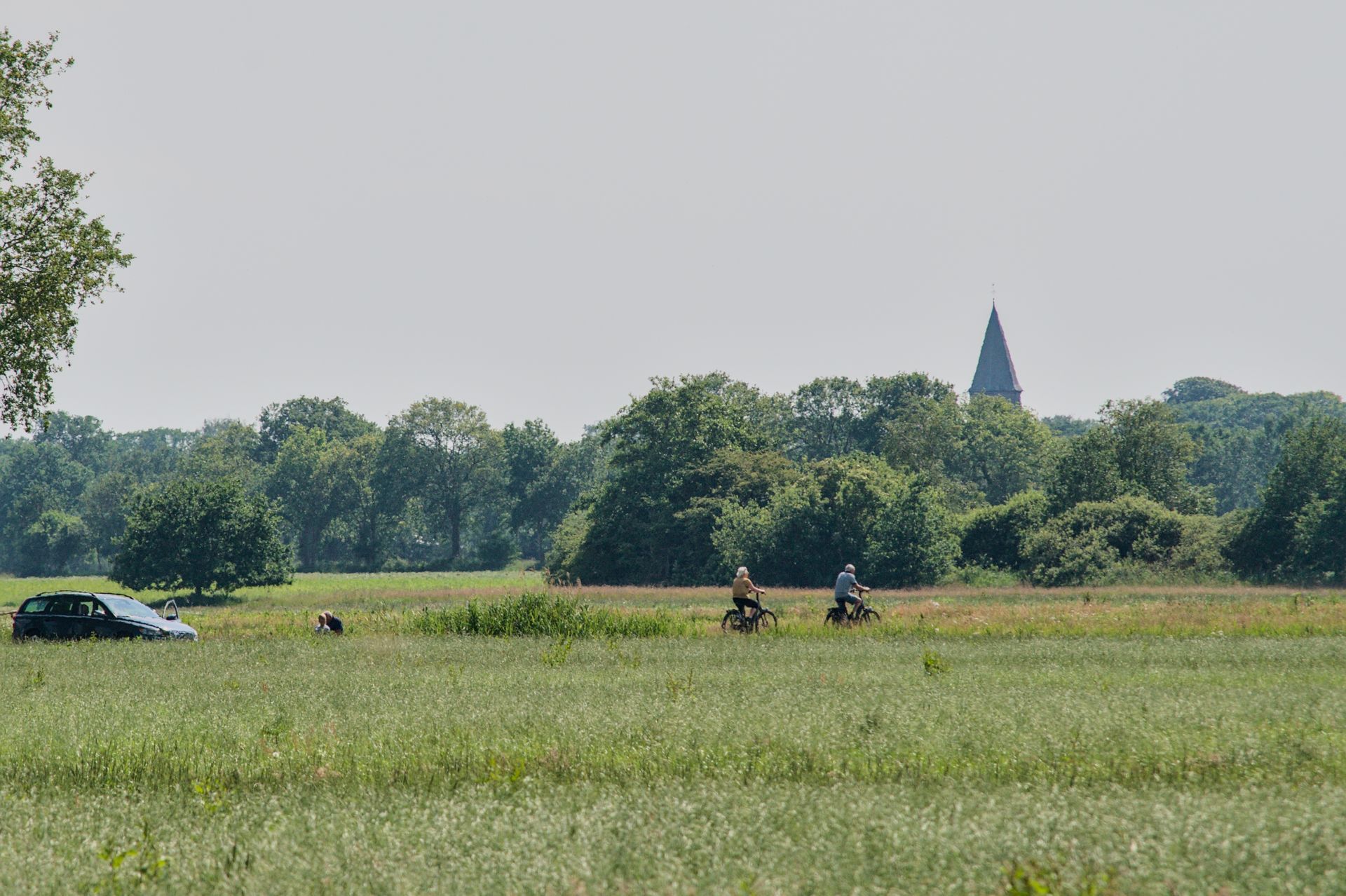 Een open veld met twee fietsers, een geparkeerde auto en bomen in de verte onder een wazige hemel.