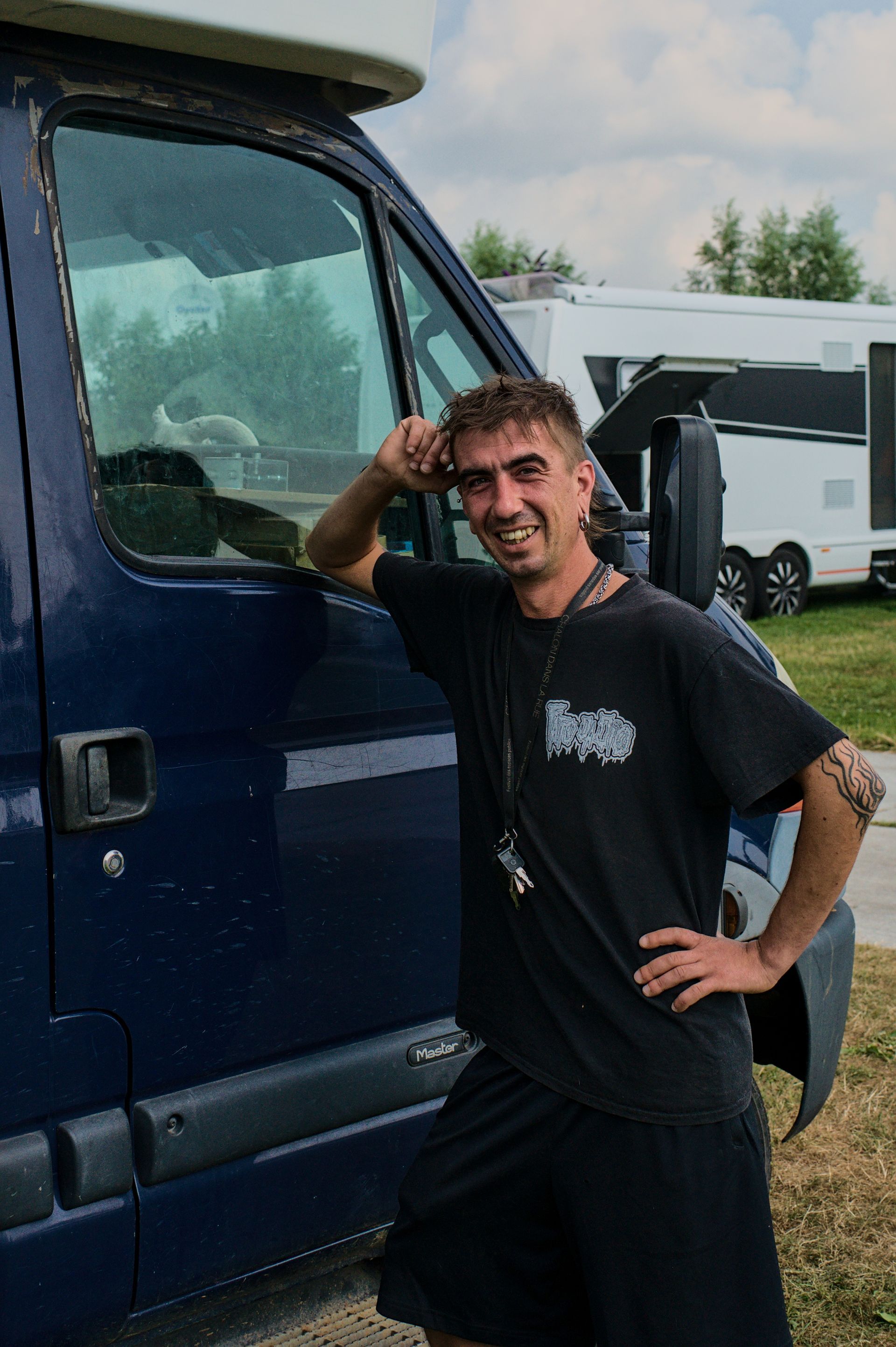 Een jonge man staat buiten naast een blauwe bestelwagen, gekleed in een zwart T-shirt en korte broek, met geparkeerde trailers op de achtergrond.