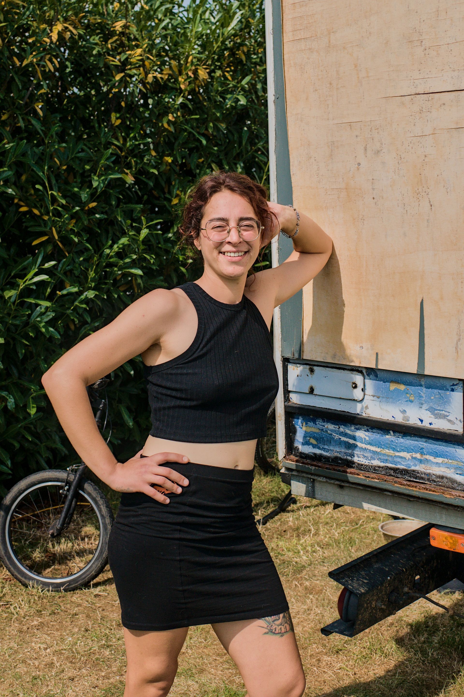 Een jonge dame poseert buiten naast een vrachtwagen en een fiets, gekleed in zwart en met een glimlach.