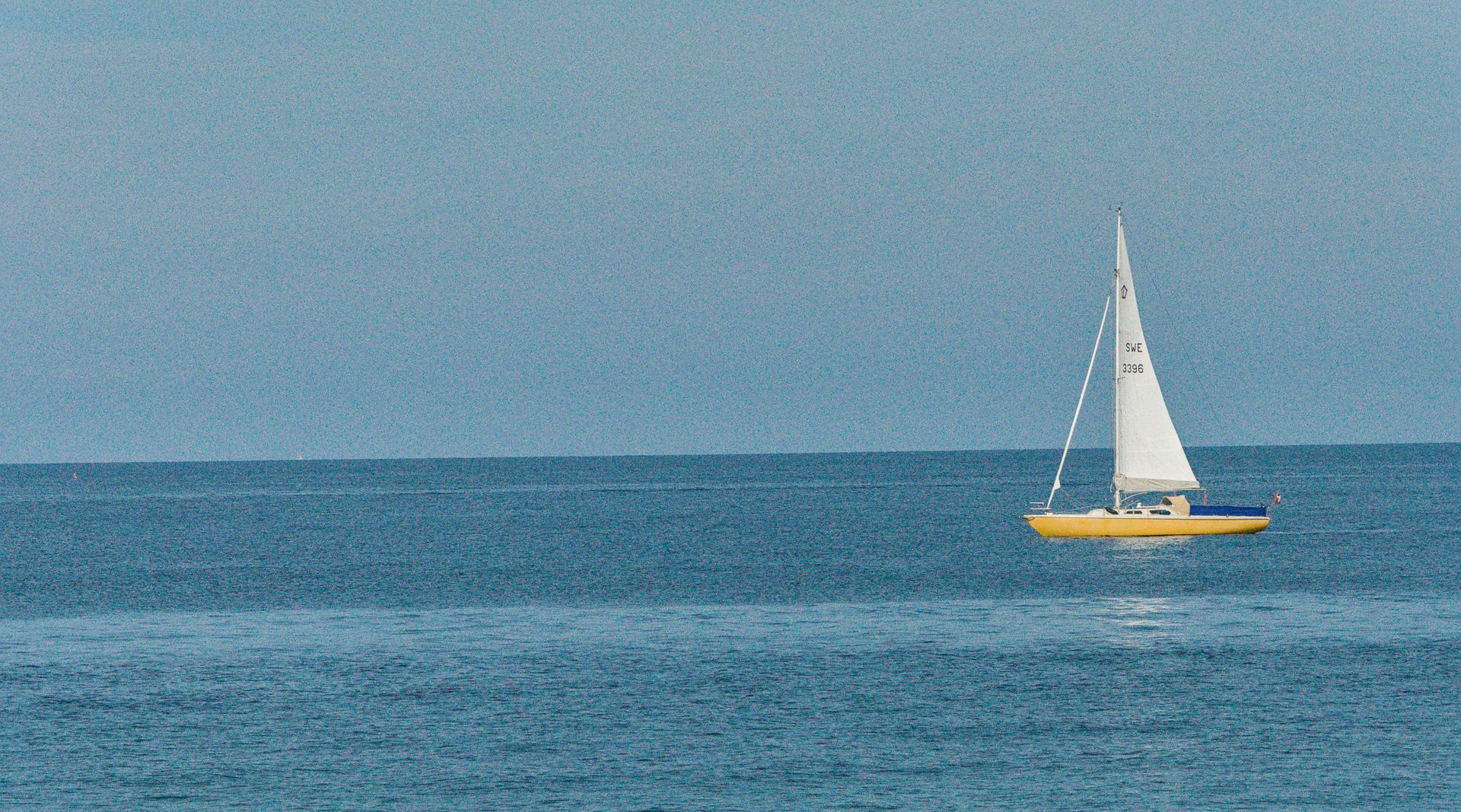 Een klein zeilbootje met een wit zeil op kalm blauw water onder een heldere hemel.
