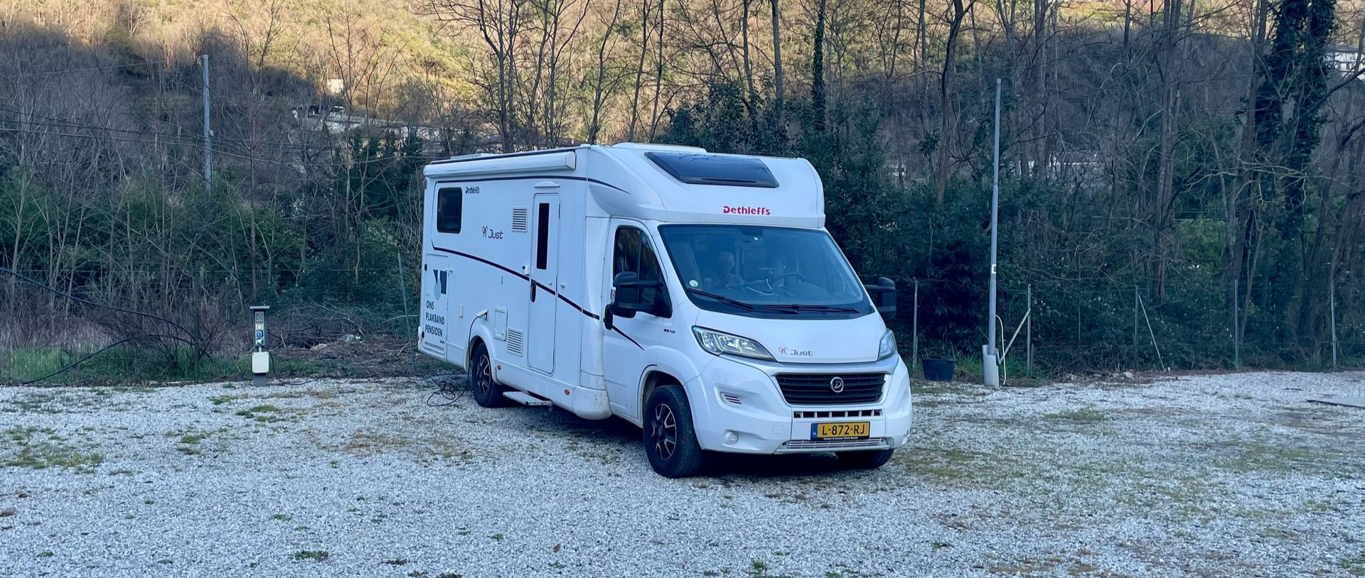 Witte camper geparkeerd op een grindterrein naast bomen en heuvels.