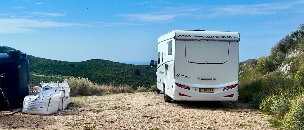 Witte camper geparkeerd op een grindterrein met de zee en groene heuvels op de achtergrond.
