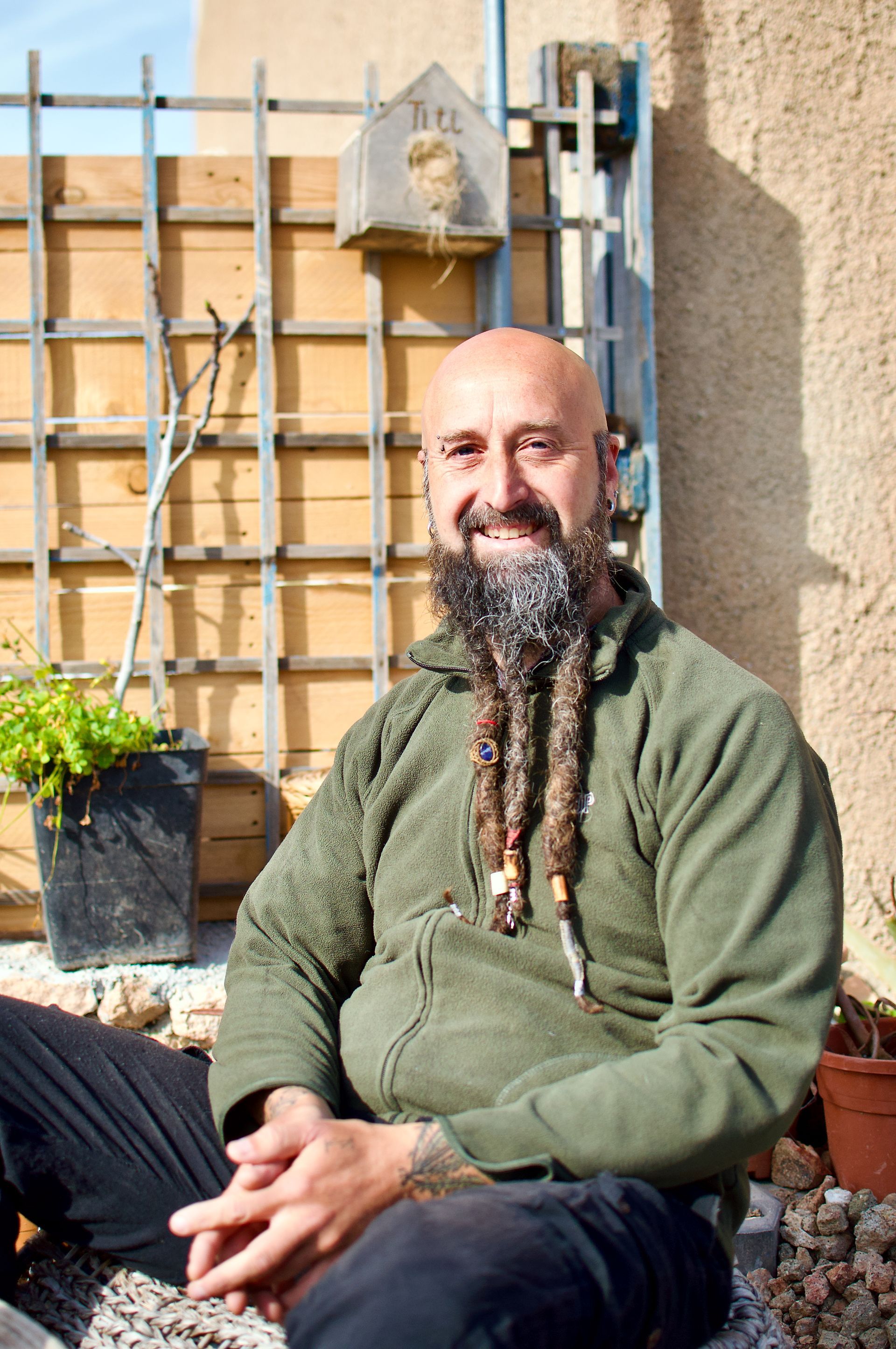 Een man met een lange baard zit buiten naast een pergola en een potplant en lacht in het zonlicht.