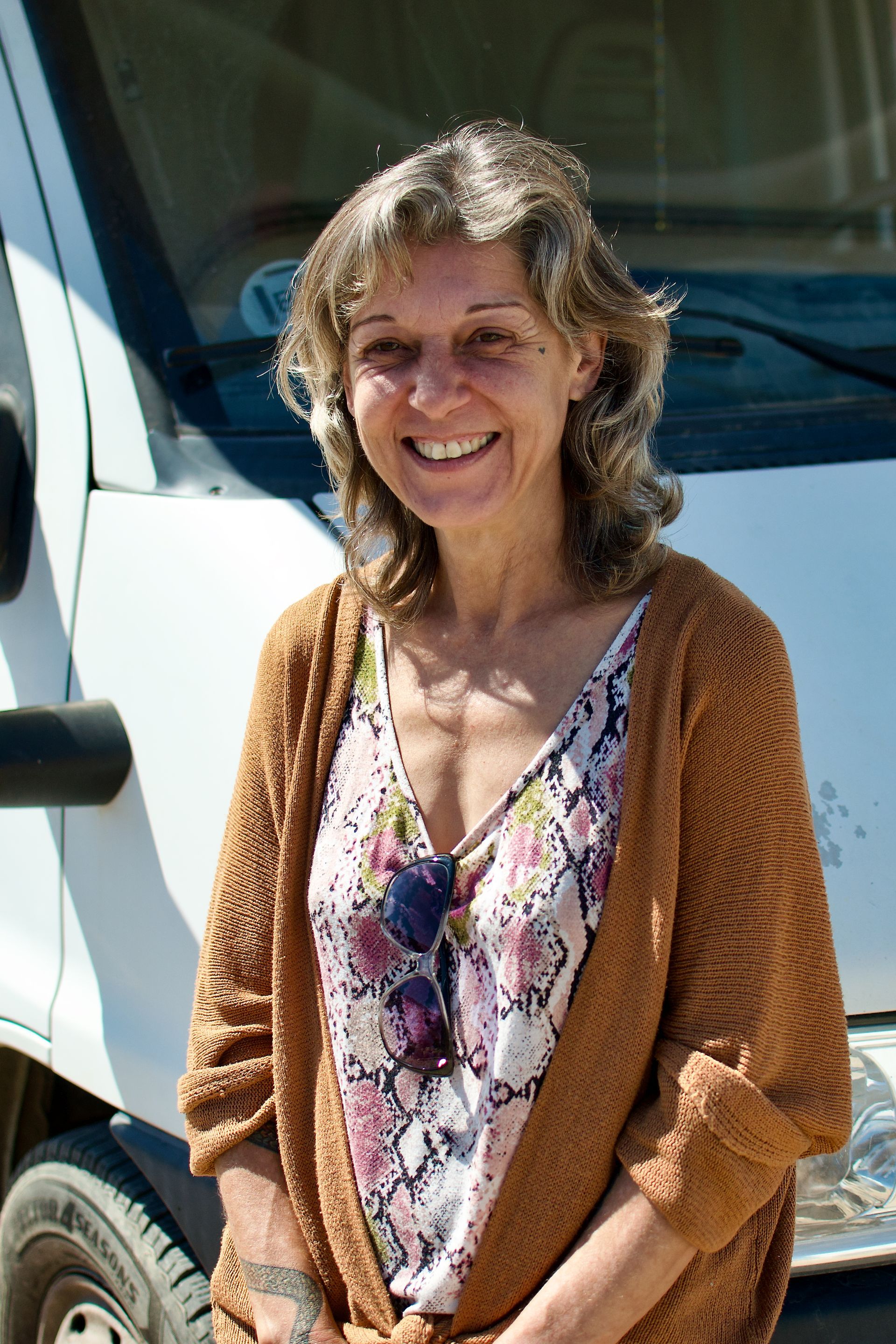 Een vrouw lacht naast een witte bestelwagen, gekleed in een top met bloemenprint en een bruin vest.