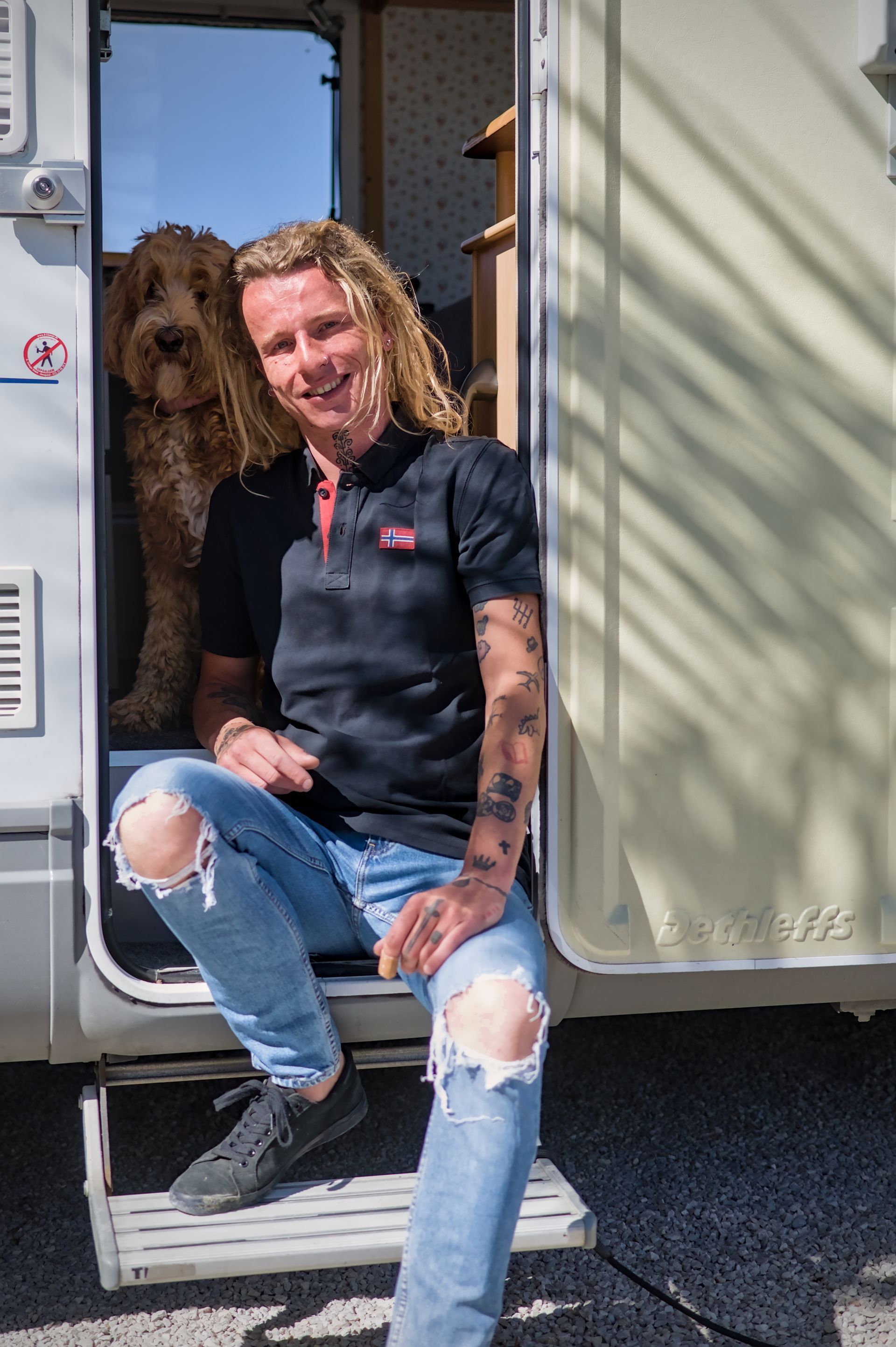 Een man zit in de deuropening van een camper, gekleed in een zwart shirt en gescheurde spijkerbroek, met een hond achter hem.