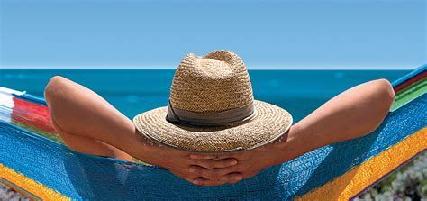 Relaxen in een hangmat Person relaxing in a hammock by the ocean, wearing a straw hat, with arms behind their head.