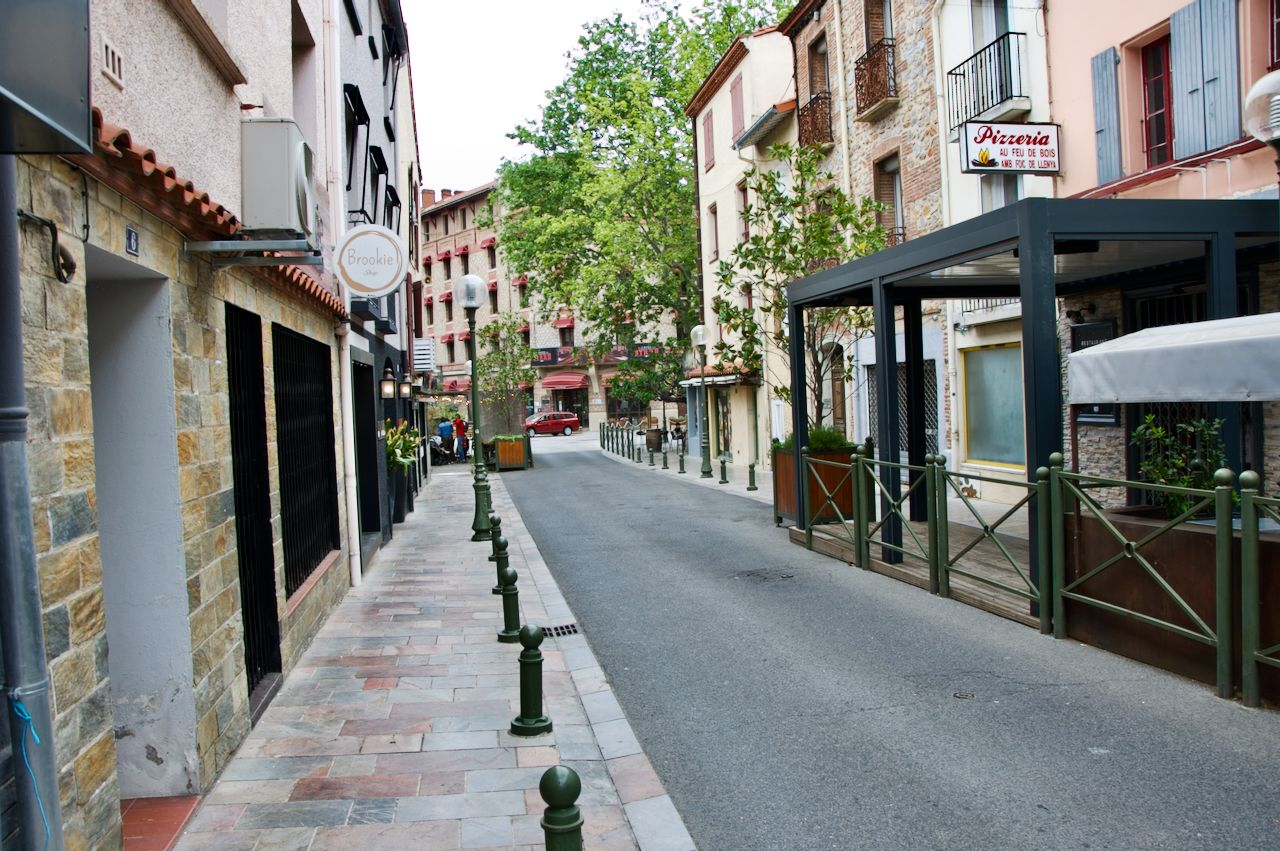 Een smalle, geplaveide straat met stenen gebouwen, terrasjes en bomen in een rustig Europees stadje.