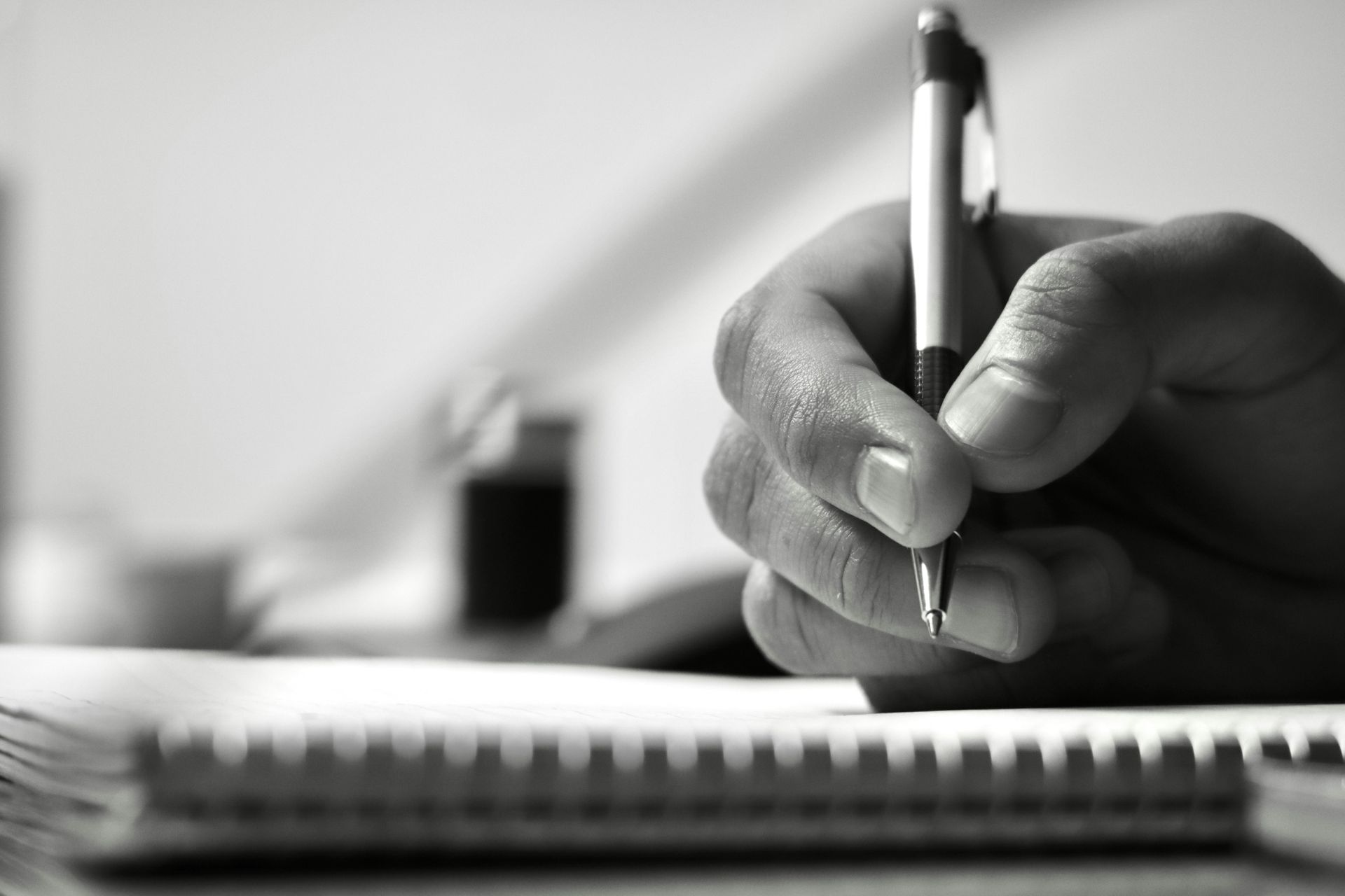 Hand holding a pen and writing in a notebook, with a blurred desk background.