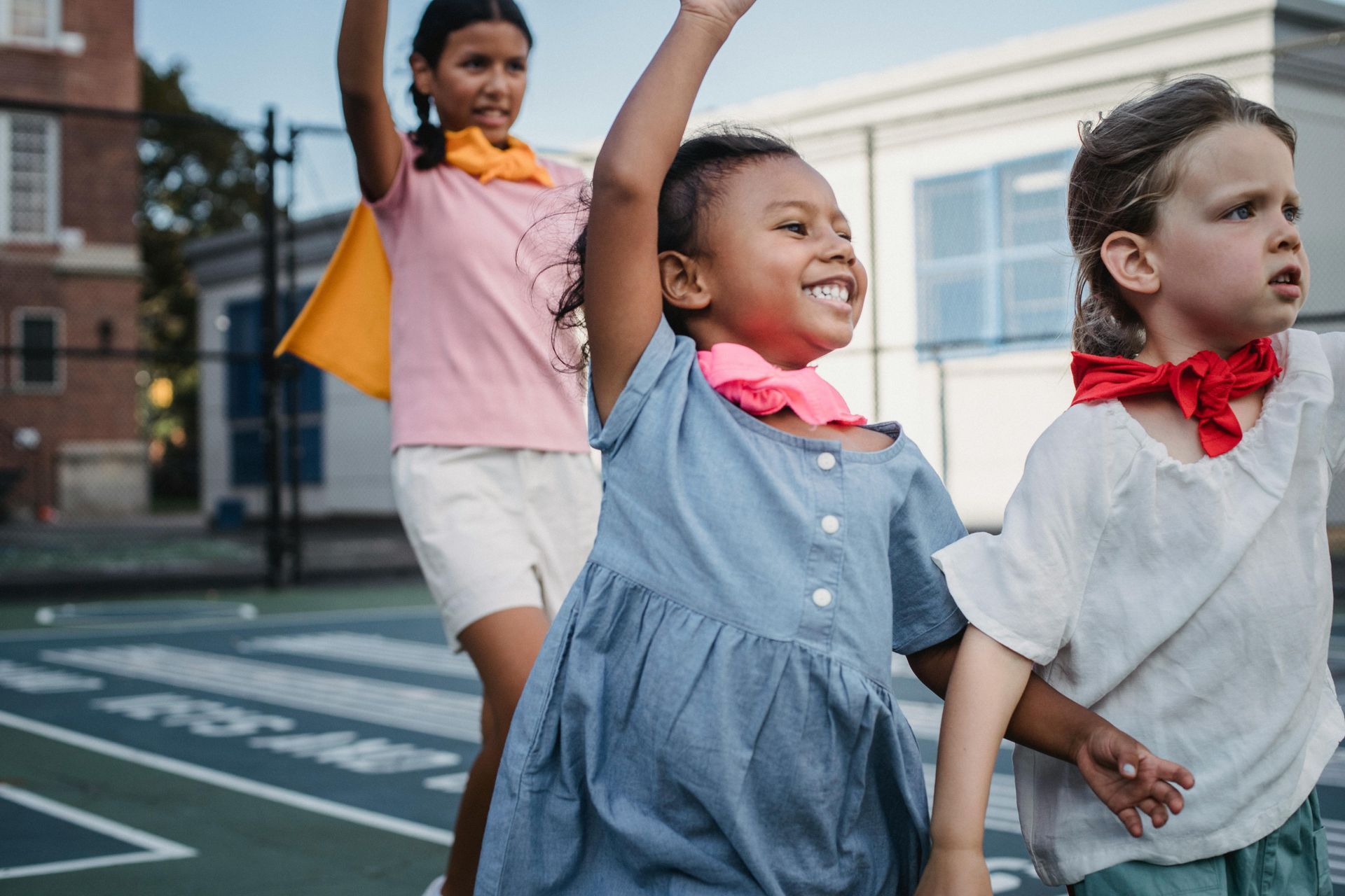 Three children holding hands and walking in a crosswalk, one smiling with an arm raised.