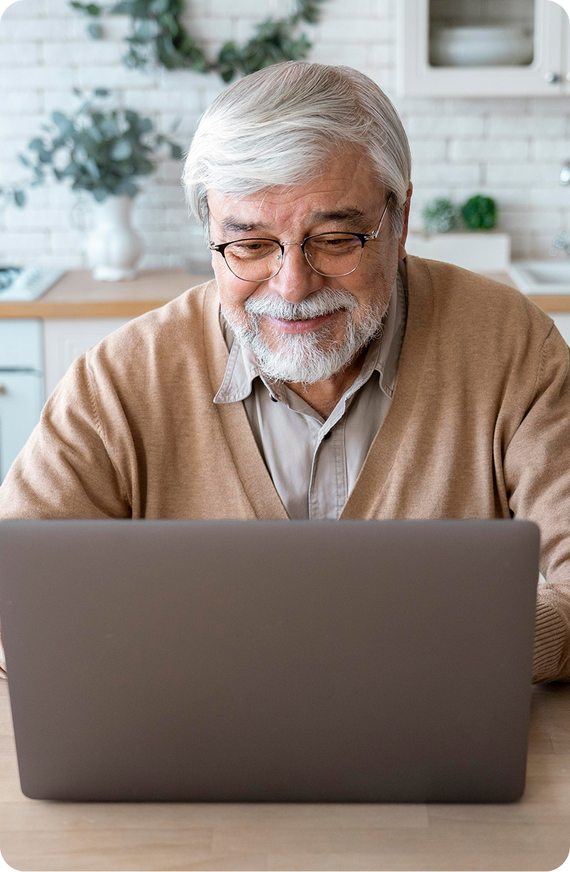 Older man smiling while looking at a laptop in a kitchen setting.