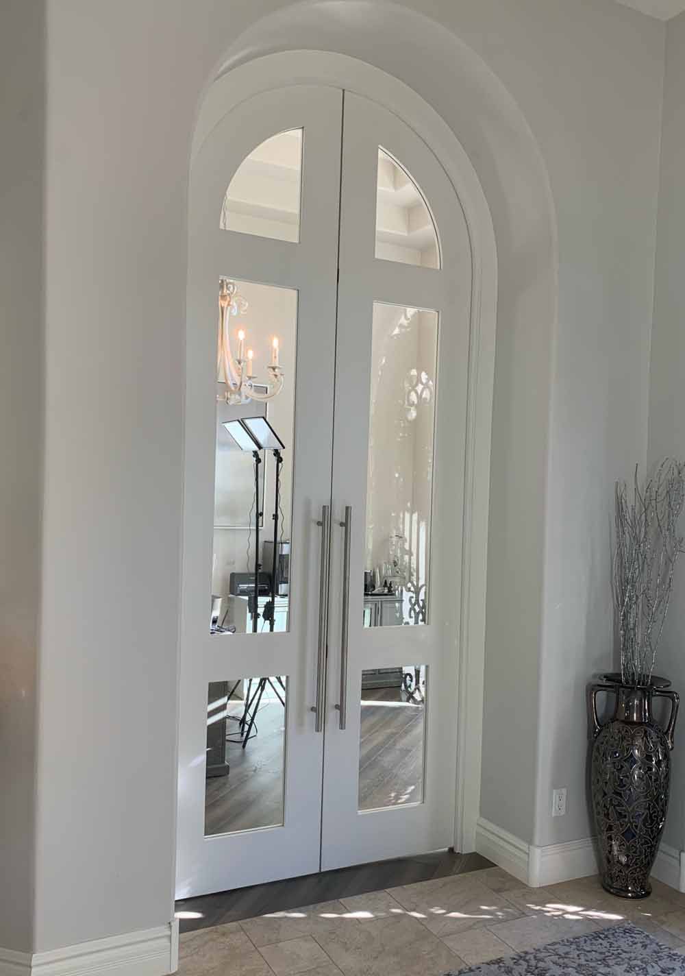 White arched double doors with glass panels and long silver handles set in a neutrally painted hallway.