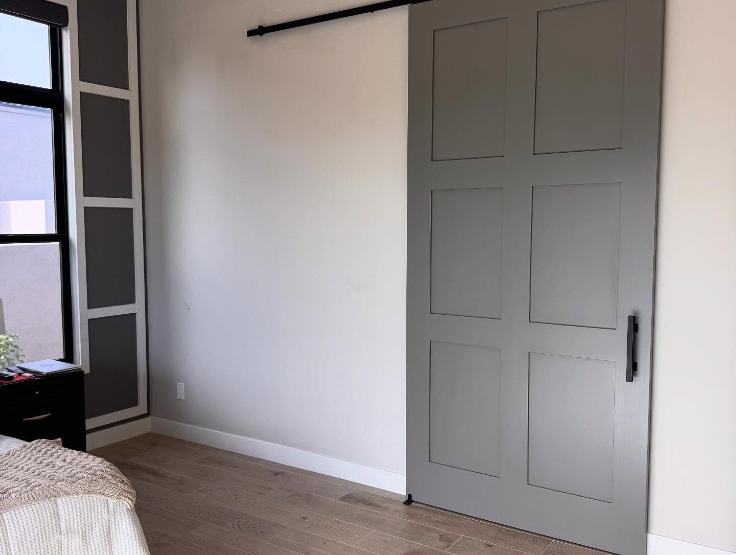 A gray six-panel sliding barn door mounted on a black rail, positioned next to a window with a dark rectangular frame.