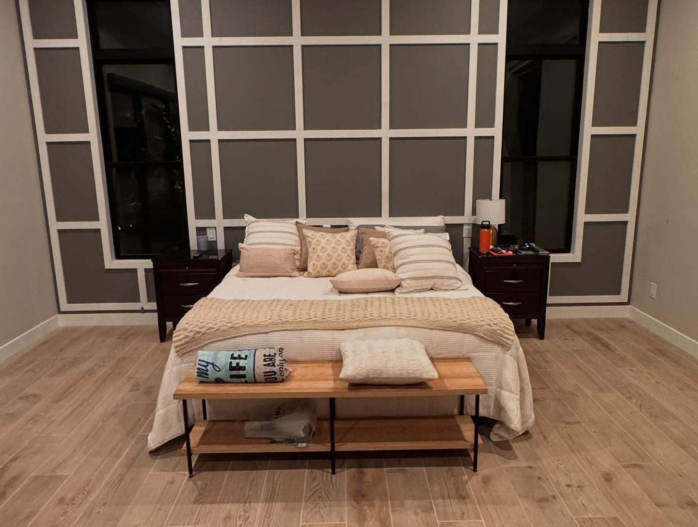 A modern bedroom with a cream-colored bed, a wooden bench, dark nightstands, and a white-grid feature wall.