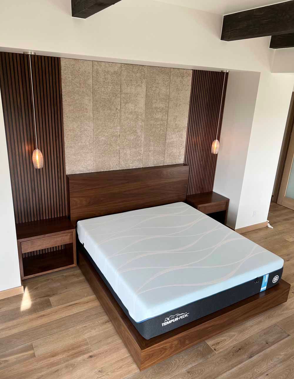 A platform bed with a grey mattress, dark wood headboard, matching nightstands, and two pendant lights against a wall.