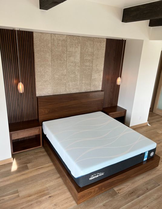 A platform bed with a gray mattress sits against a textured stone wall framed by dark wood panels and hanging lights.
