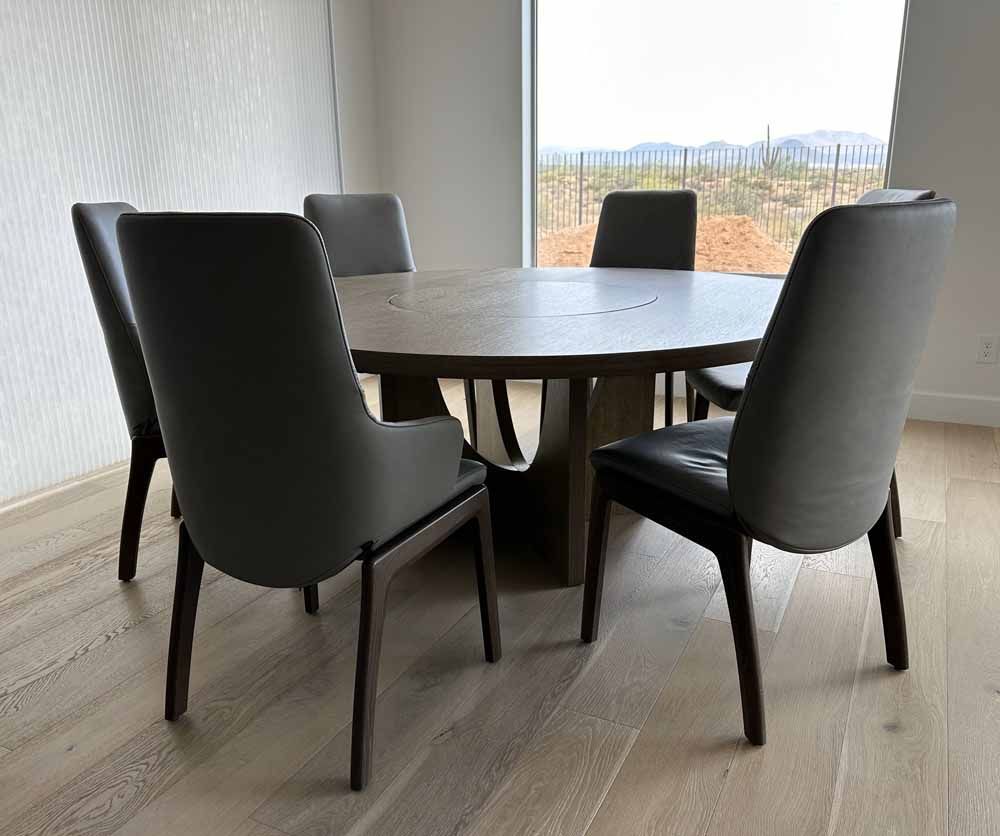 A round wooden dining table surrounded by six gray upholstered chairs in a sunlit room with light-colored flooring.