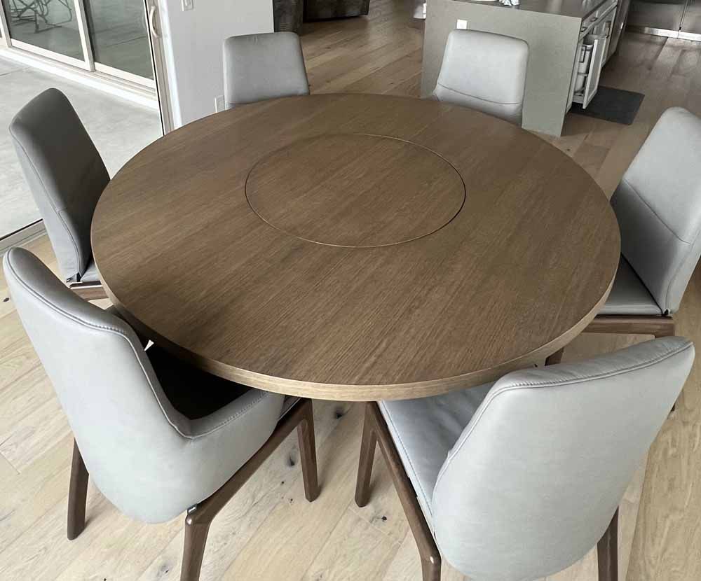 A round, light brown wooden dining table with a built-in lazy susan, surrounded by six light grey upholstered chairs.