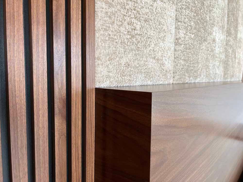 Close-up of vertical walnut wood slats next to a smooth walnut shelf against a textured, light-colored stone wall.