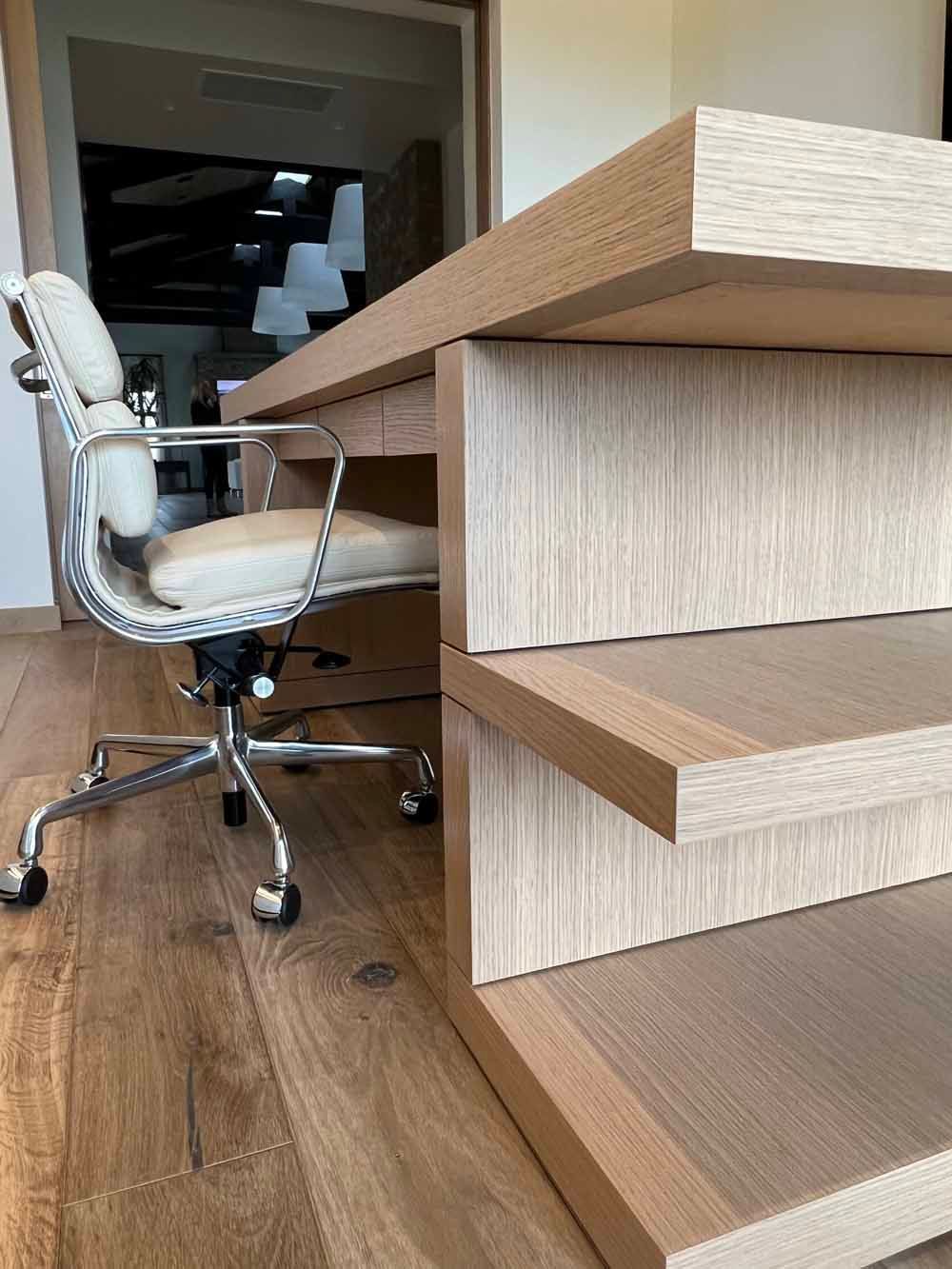 A sleek, light-wood desk with layered shelves sits next to a modern swivel office chair with white cushions.